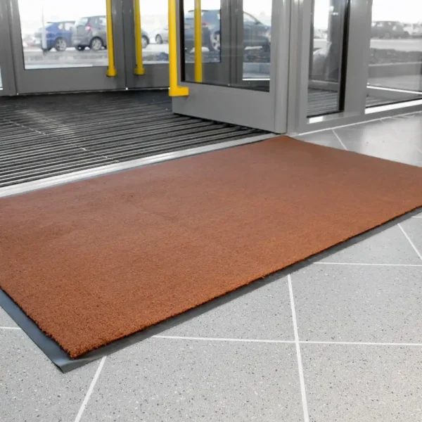 A Plush Entrance Mat in rectangular brown design is placed on a tiled floor near glass entrance doors, with cars visible outside.