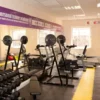 A bright gym room with weight benches, weights, a treadmill, and other exercise gear on a yellow and grey Circular Stud Interlocking Tile floor, with large mirrors and windows allowing in natural light.