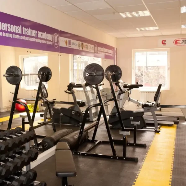 A bright gym room with weight benches, weights, a treadmill, and other exercise gear on a yellow and grey Circular Stud Interlocking Tile floor, with large mirrors and windows allowing in natural light.
