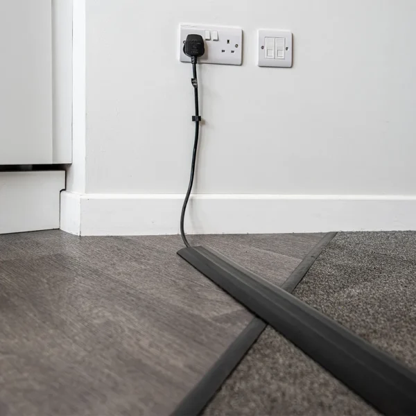 A black cable runs from a wall socket, shielded by the PVC Cable Protector as it crosses from hard floor to carpet in a room corner.