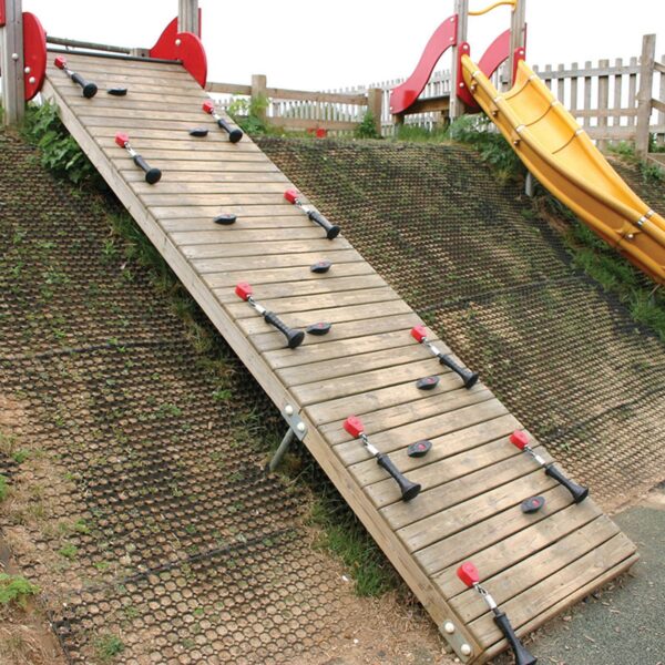 A wooden climbing ramp with hand and foot holds stands in an outdoor playground next to a bright yellow slide, with Grass Mat Roll (1m x 10m x 22mm) used for safety surfacing on the ground.
