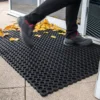 A person in black trousers and shoes steps onto a Roof Walkway Mat with circular holes, placed outside a building entrance. Yellow leaves are scattered across the ground and mat.