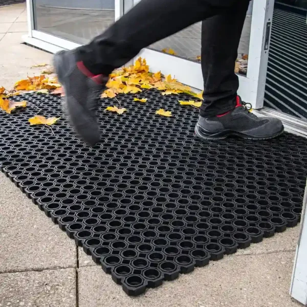 A person in black trousers and shoes steps onto a Roof Walkway Mat with circular holes, placed outside a building entrance. Yellow leaves are scattered across the ground and mat.