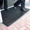 Two people in dark trousers and dress shoes walk across a Drainage Top Mat with circular holes, placed at a building entrance with glass doors and brick paving outside.