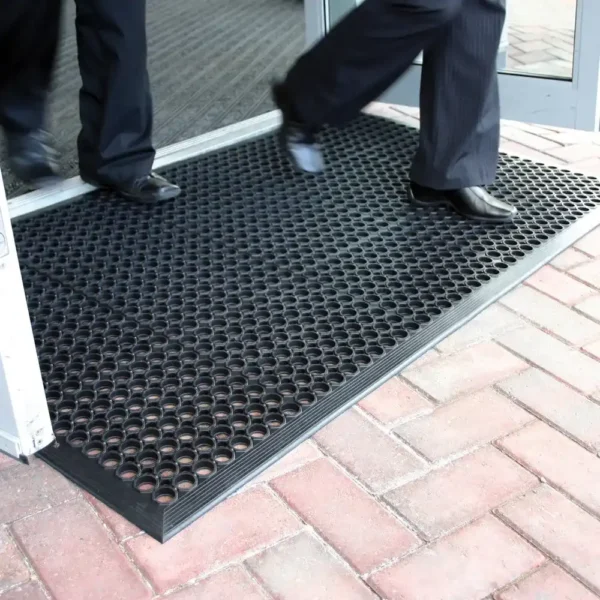 Two people in dress trousers and shoes step onto a Drainage Top Mat, a black rubber doormat with circular holes, placed at the entrance of a building with brick paving outside.