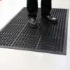 A person in black shoes stands on a Drainage Top Mat, a black rubber anti-fatigue mat with circular holes, placed on a light-coloured floor.