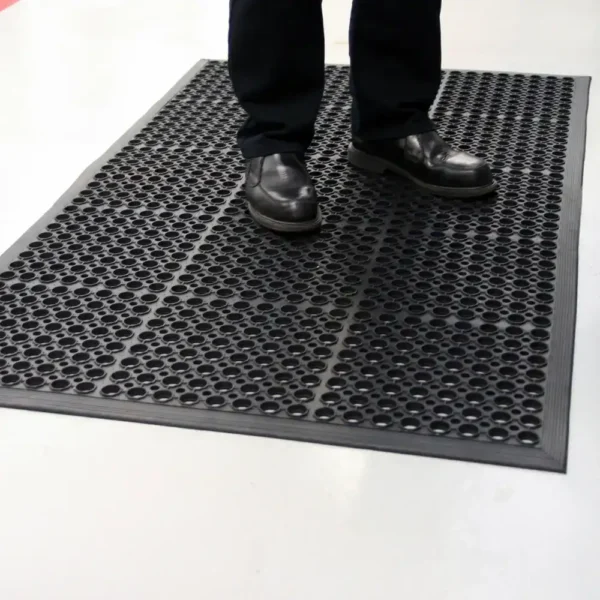 A person in black shoes stands on a Drainage Top Mat, a black rubber anti-fatigue mat with circular holes, placed on a light-coloured floor.