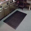 A Drainage Top Mat—black anti-fatigue rubber with circular holes—is placed on the floor in front of a stainless steel kitchen counter and cooker in a commercial kitchen.