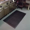 A black Drainage Top Mat with holes is placed on the grey floor in front of stainless steel kitchen appliances and a metal prep table in a commercial kitchen.