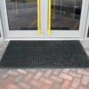 The Large Front Door Mat Bundle, a black rubber mat with holes, is set before a glass double door with yellow handles on interlocking brick paving.