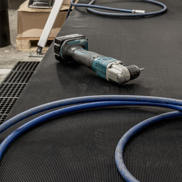 A cordless power drill and a blue air hose rest on Fine Ribbed Rubber Matting in a workshop, with cardboard boxes and other tools visible in the background.