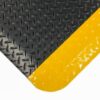 A close-up of the Deckplate Anti-Fatigue Mat shows its textured, glossy black surface with a diagonal diamond pattern for grip and a bold yellow stripe across the corner.