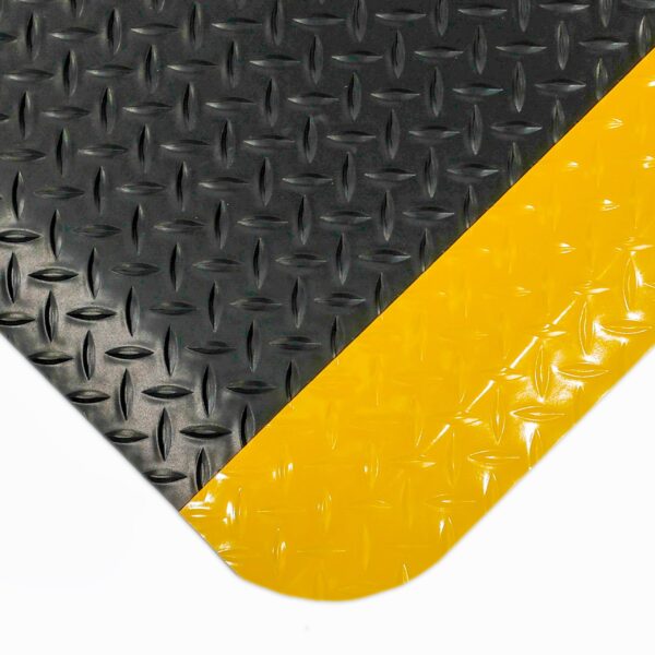A close-up of the Deckplate Anti-Fatigue Mat shows its textured, glossy black surface with a diagonal diamond pattern for grip and a bold yellow stripe across the corner.