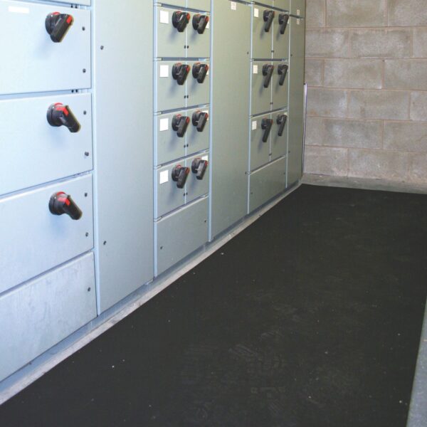 A room with grey electrical switchgear cabinets featuring black and red handles lines the left wall, while Electrical Insulation Matting is placed on the floor beside the cabinets. The walls are constructed of breeze blocks.