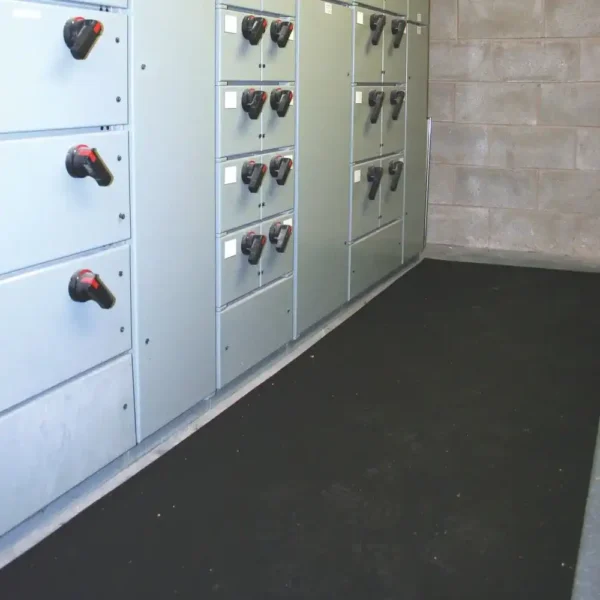 A black Electrical Insulation Matting is placed on the floor in front of electrical control panels with switches in an industrial setting with concrete block walls.