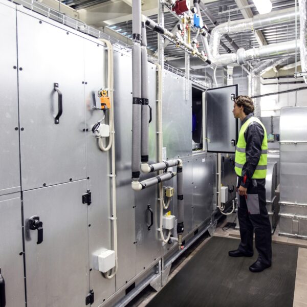 A person wearing a safety vest and work clothes checks industrial equipment inside a plant room with shiny metal pipes and machinery, standing on Electrical Safety Matting for protection.