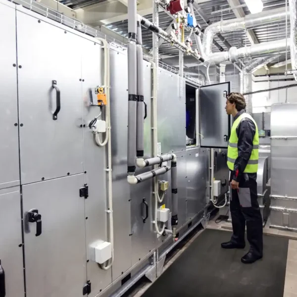 Wearing safety gear and a high-visibility vest, a person inspects large industrial HVAC equipment inside a facility with metal pipes and ducts, whilst standing on Electrical Safety Matting for added protection.