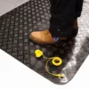 A person in black trousers and brown safety boots stands on a Senso Dial Mat ESD. A yellow earthing cable with a clip and plug is placed on the mat near their feet.