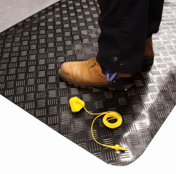 A person in black trousers and brown safety boots stands on a Senso Dial Mat ESD. A yellow earthing cable with a clip and plug is placed on the mat near their feet.