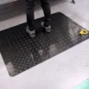 A person stands on a Senso Runner ESD black anti-fatigue mat with a chequered pattern in a workspace; a yellow tape measure is on the floor nearby.