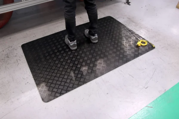 A person stands on a Senso Runner ESD black anti-fatigue mat with a chequered pattern in a workspace; a yellow tape measure is on the floor nearby.