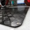 Close-up of the Senso Runner ESD, a black textured anti-slip floor mat with a metal press stud; a person in trainers stands on it, blurred in the background.