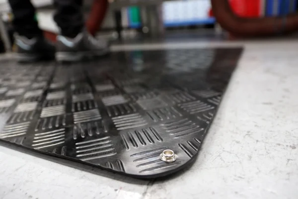 Close-up of the Senso Runner ESD, a black textured anti-slip floor mat with a metal press stud; a person in trainers stands on it, blurred in the background.