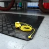 A pair of yellow Senso Runner ESD earplugs, connected by a cord, rests on a black textured rubber mat placed on a grey industrial floor. Equipment and red hoses can be seen in the background.