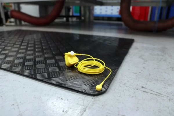A pair of yellow Senso Runner ESD earplugs, connected by a cord, rests on a black textured rubber mat placed on a grey industrial floor. Equipment and red hoses can be seen in the background.