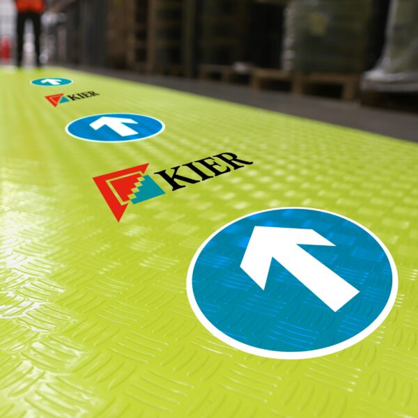 The Construction Site Walkway Roll is a green floor covering with blue circular signs featuring white arrows and the KIER logo, used to indicate walking direction in indoor industrial settings.