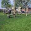 Children play on wooden climbing and swinging equipment installed on a Grass Mat Roll (1m x 10m x 22mm) in a grassy outdoor playground surrounded by trees and houses on a sunny day.