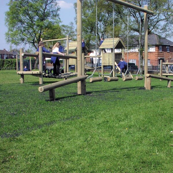 Children play on wooden climbing and swinging equipment installed on a Grass Mat Roll (1m x 10m x 22mm) in a grassy outdoor playground surrounded by trees and houses on a sunny day.