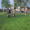 Children play on grass protected by the 16mm Black Grass Mat, climbing a wooden frame and swinging in a sunny playground bordered by trees and houses.
