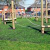 A Grass Mat Roll (1m x 10m x 22mm) is installed beneath wooden playground climbing equipment with chains and ropes on a sunny park lawn, with houses and trees visible in the background.