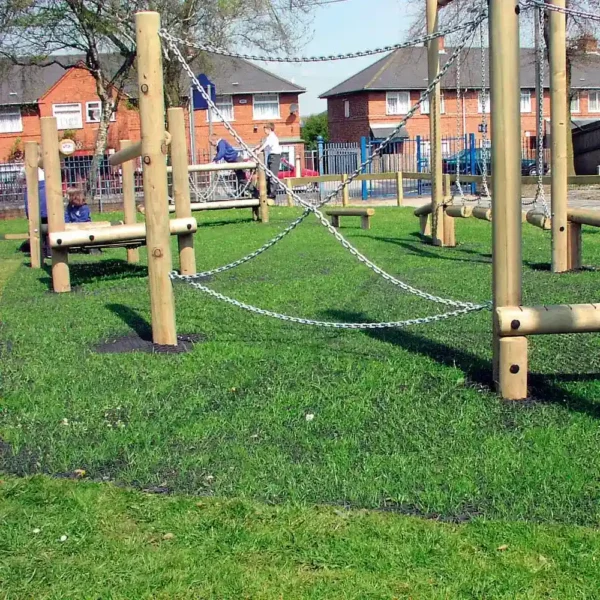 A wooden climbing frame with chains is installed on 16mm Black Grass Mat artificial turf, enclosed by a metal fence. Houses and trees can be seen in the sunny background.