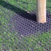 A wooden post stands in grass atop the Grass Mat Roll - 1m x 10m x 22mm, featuring black rubber with a honeycomb pattern that lets grass grow through the holes.