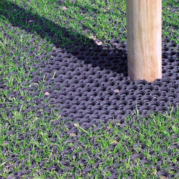 A wooden post stands in grass atop the Grass Mat Roll - 1m x 10m x 22mm, featuring black rubber with a honeycomb pattern that lets grass grow through the holes.
