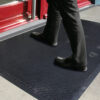 A person in black dress trousers and shoes steps onto the Dirt Remover Mat, a textured black mat placed on a grey surface by a red door entrance.