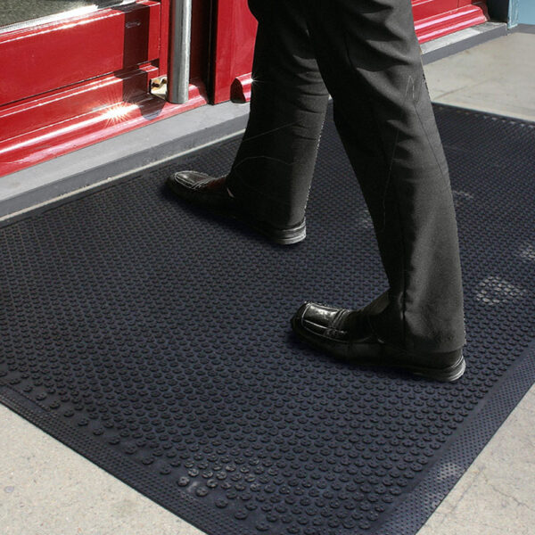 A person in black dress trousers and shoes steps onto the Dirt Remover Mat, a textured black mat placed on a grey surface by a red door entrance.