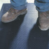 Close-up of a person in brown work boots and blue trousers standing on the Dirt Remover Mat, a black textured rubber mat featuring a slightly raised circular pattern.