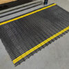 The Self-Draining Safety Mat, featuring yellow stripes, is placed on a grey industrial floor in front of shelving to prevent slipping and clearly mark a safe work zone.