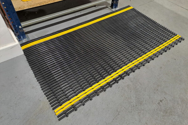 The Self-Draining Safety Mat, featuring yellow stripes, is placed on a grey industrial floor in front of shelving to prevent slipping and clearly mark a safe work zone.