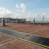 A flat rooftop features brown gravel and Self-Draining Safety Mats, edged by metal railings. Yellow and white cables cross the surface, while distant buildings and water are visible beneath a partly cloudy sky.