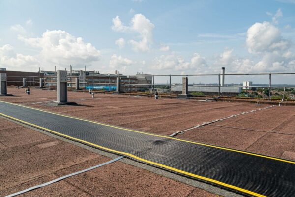 A flat rooftop features brown gravel and Self-Draining Safety Mats, edged by metal railings. Yellow and white cables cross the surface, while distant buildings and water are visible beneath a partly cloudy sky.