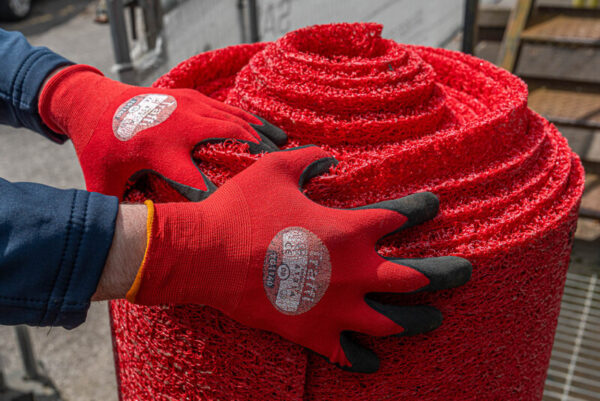 A person wearing red gloves with black fingertips is handling a large roll of Heavy-Duty PVC Vinyl outdoors, possibly during installation or transport.