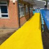 A bright yellow Heavy-Duty PVC Vinyl Roll forms a pathway along a brick building with large windows, bordered by blue safety barriers. Two people are visible in the distance near the end of the path.