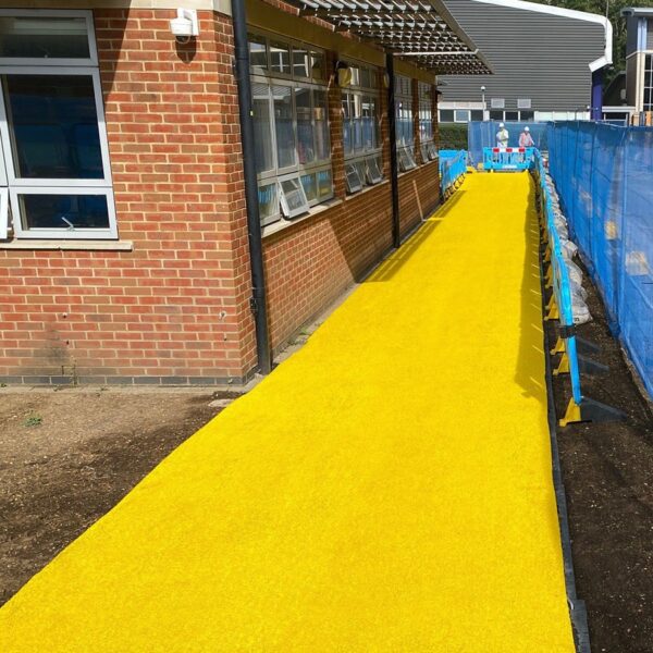 A bright yellow Heavy-Duty PVC Vinyl Roll forms a pathway along a brick building with large windows, bordered by blue safety barriers. Two people are visible in the distance near the end of the path.