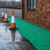 Three orange and white traffic cones are set on a wet surface beside a Heavy-Duty PVC Vinyl Roll, placed along a brick building, with gravelly ground and a wooden fence visible in the background.