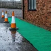 Three orange and white traffic cones are set along the edge of a green Heavy-Duty PVC Vinyl Roll near a brick building on wet gravel, with a wooden fence visible in the background.