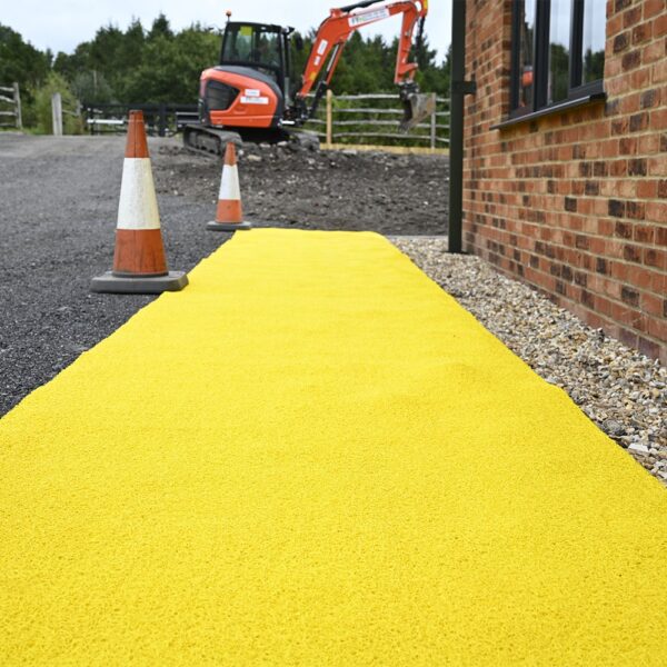 A bright yellow Heavy-Duty PVC Vinyl Roll forms a textured pathway alongside a brick building, with two orange traffic cones and an orange digger seen in the gravel area behind.